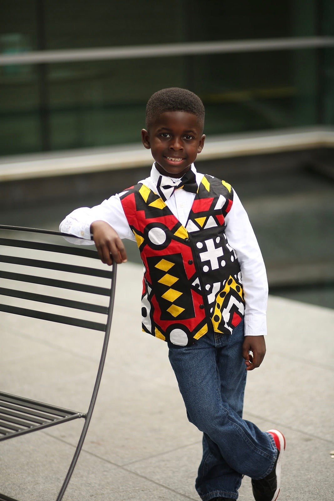 African American boy leaning on a bench and wearing an African Ankara print of red, black, yellow and white with geo,ethic shapes. The vest has black snaps or black buttons. Vest comes with coordinating bow tie of the same fabric.