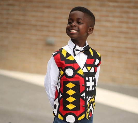 African American boy smiling while wearing an African Ankara print of red, black, yellow and white with geo,ethic shapes. The vest has black snaps or black buttons. Vest comes with coordinating bow tie of the same fabric.