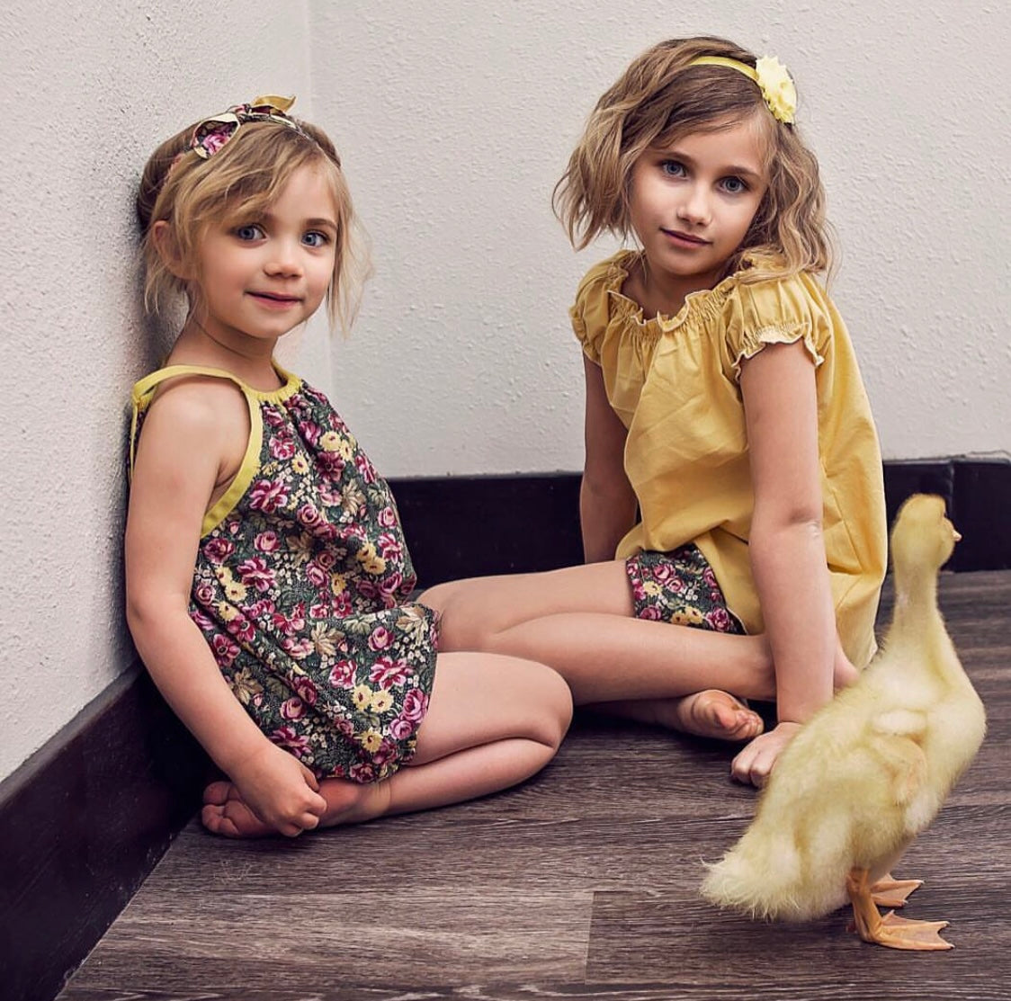Sisters wearing coordinating outfits from Ackee Tree Clothing. A sweet dark floral baby bubble romper with yellow trim and matching knotted head tie. And a buttercup yellow boho blouse with shorted neckline and the coordinating dark floral shorts.
