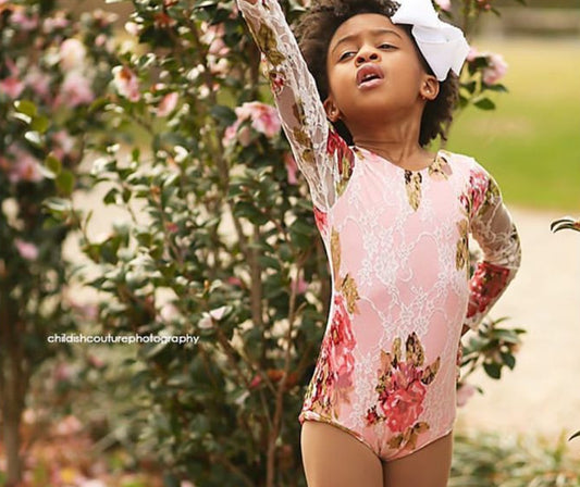 Little African American girl wearing a lace leotard that is lined with a soft stretch fabric. The lace is ivory with reddish pink and green florals. 