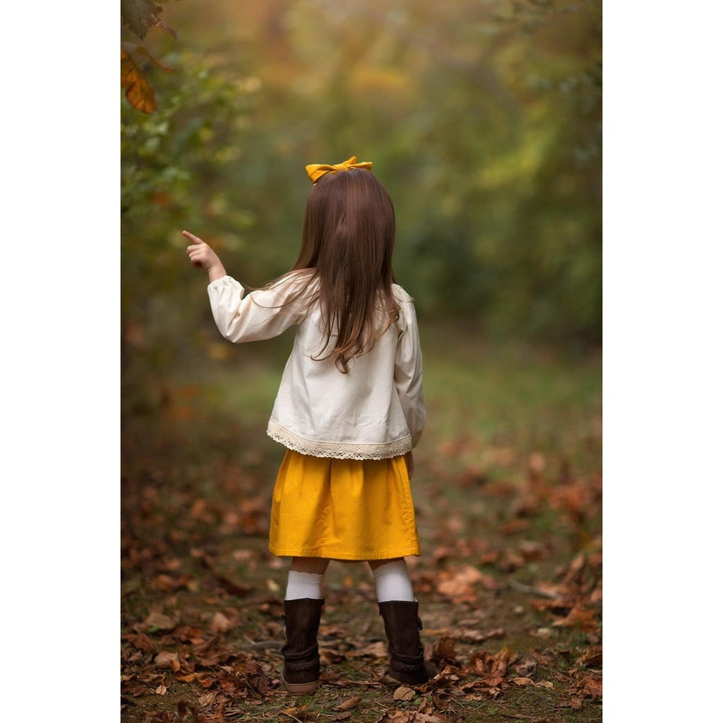 Back view of a Little girl wearing a boho skirt set with a mustard yellow skirt and hair accessory and an Ecru colored peasant top with lace trim.