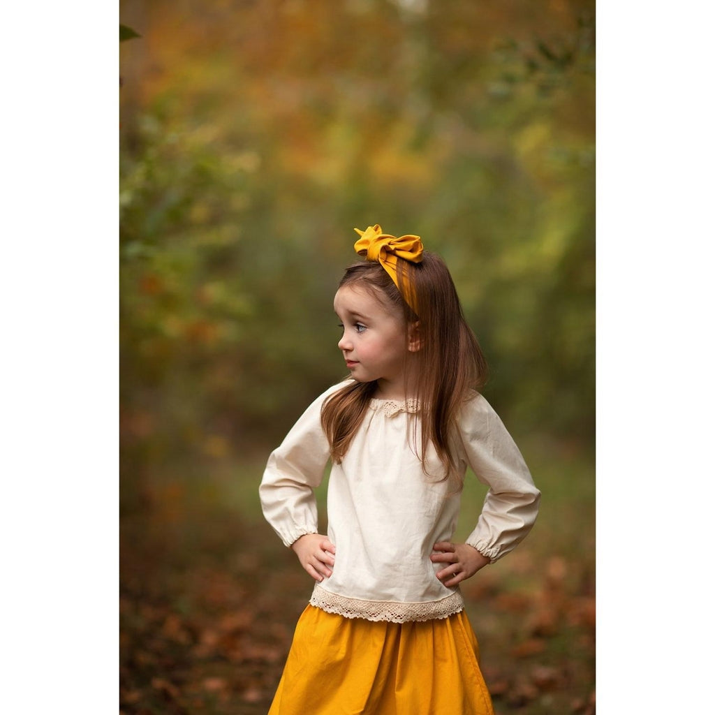 Little girl wearing a boho skirt set with a mustard yellow skirt and hair accessory and an Ecru colored peasant top with lace trim.