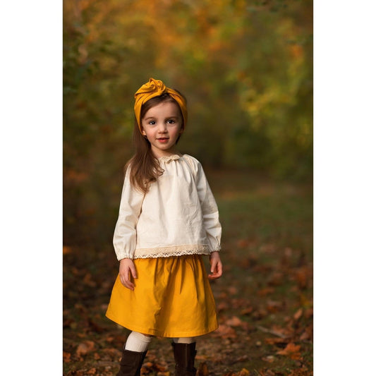 Little girl wearing a boho skirt set with a mustard yellow skirt and hair accessory and an Ecru colored peasant top with lace trim.